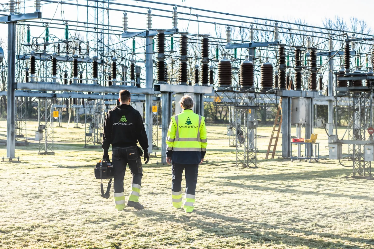 Two technicians in personal protective equipment inspect an HV/MV transformer substation at dawn